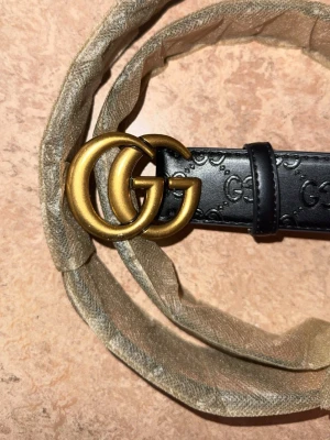 Svart Gucci skärp med GG-spänne - Snyggt svart skärp från Gucci med präglade GG-loggor längs hela bandet och ett stort, guldfärgat GG-spänne framtill. Skärpet är gjort i skinn och har en klassisk, tidlös design som lyfter vilken outfit som helst. 