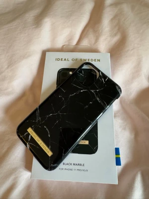 Ideal Of Sweden Svart marmormönstrat mobilskal - Snyggt och enkelt svart mobilskal med marmormönster från iDeal of Sweden. Skalet har en elegant finish med vita ådror och en guldfärgad logotyp. Passar iPhone 11 Pro/XS/X och är tillverkat i hårt material som skyddar mobilen bra! Så snygg😻Använd några gånger men inga fel🩷Orginal pris runt 399kr.