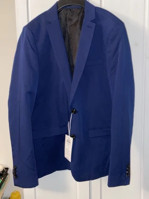 Blå kavaj från Jack & Jones - Snygg blå kavaj från Jack & Jones i modellen JPRSOLA BLAZER NOOS JNR. Kavajen har två knappar framtill, klassisk krage och två fickor. Fodrad insida och knappdetaljer vid ärmslut. Perfekt för en stilren look.