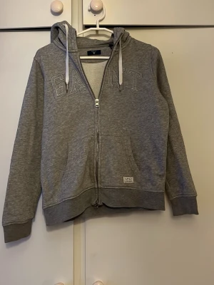 Grå zip-hoodie från GANT - Säljer en grå zip-hoodie från GANT med broderad logga på bröstet och två fickor framtill. Tröjan har huva med vita snören och ribbade muddar vid ärmslut och nederkant. Perfekt för en avslappnad och cool look.