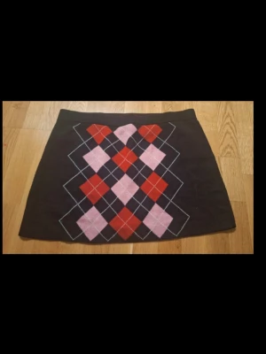 Rutig svart stickad kortkjol med argylemönster - Snygg svart kortkjol med klassiskt argylemönster i rött och rosa framtill. Kjolen har en rak passform och är perfekt för dig som vill sticka ut med en färgglad detalj. Passar till både sneakers och boots för en trendig look.