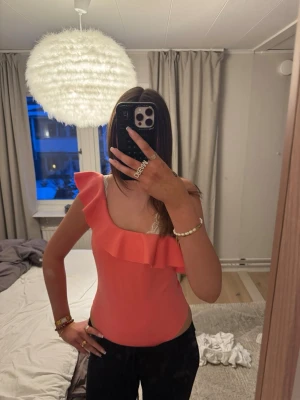 Korallrosa body med volang - Snygg korallrosa body med en axel och volangdetalj längs halsringningen. Tight passform och stretchigt material som sitter bekvämt. Perfekt för dig som vill ha en trendig och feminin look.