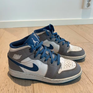 Nike Air Jordan 1 Mid grå/blå sneakers - Säljer ett par Nike Air Jordan 1 Mid sneakers i grått, vitt och blått. Klassisk high-top modell med blå snörning, ikonisk Swoosh och Air Jordan-logga på sidan. Skorna har en sportig vibe och är perfekta för dig som gillar streetwear.
