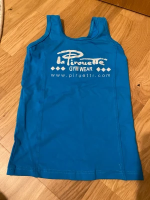 Blått linne från Piruetti Gym Wear - Snyggt blått träningslinne från Piruetti Gym Wear med tryck på bröstet. Linnet har breda axelband och är tillverkat i ett stretchigt och mjukt material som passar perfekt för gym eller dans. Enkel och sportig design. Linnet är dok använd rikligt med gånger så en liten tråd saknas men syns inte när man använder den!💓