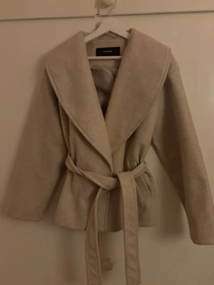 Beige kappa med knytskärp - Stilren beige kappa med bred krage och knytskärp i midjan. Kappan har en avslappnad passform och är tillverkad i ett mjukt ull-liknande material. Perfekt för att hålla värmen och se trendig ut under kyliga dagar.