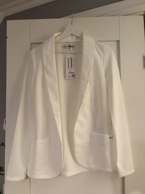 Vit kavaj från New Yorker med fickor - Stilren vit kavaj/blazer från New Yorker med två framfickor och klassisk krage. Rak passform och utan knäppning framtill. Perfekt för en clean och modern look. Materialet är mjukt och har en lätt stretch för extra komfort.