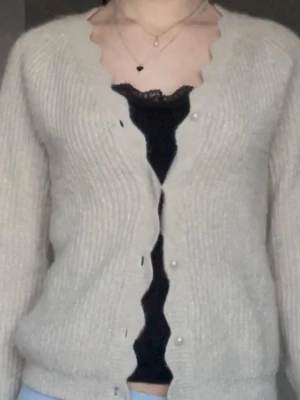 Beige kofta med vågig kant från Vila - Mysig beige stickad kofta från Vila med vågig kant längs knäppningen och pärlliknande knappar. Koftan har v-ringning, lång ärm och ribbstickad struktur. Perfekt att slänga över ett linne eller t-shirt för en chill look. OBS, saknar en pärlknapp längst ner! 