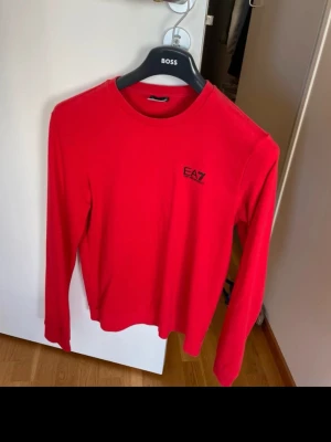 Röd tröja från EA7 - Snygg röd långärmad tröja från EA7 med rund halsringning och svart EA7-logga på bröstet. Tröjan har en enkel och stilren design med svarta ränder i nacken på baksidan. Perfekt för dig som gillar sportig och modern stil.