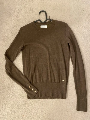 Brun stickad tröja från Holly & Whyte - Snygg brun stickad tröja från Holly & Whyte med rund halsringning och långa ärmar. Tröjan har ribbade muddar och dekorativa guldfärgade knappar på ärmsluten. Materialet är mjukt och tunt, perfekt för lager-på-lager. Diskret logotyp i metall på framsidan.