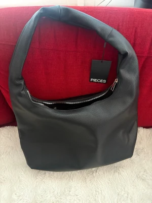 Svart axelväska från Pieces - Stilren svart axelväska från Pieces i mjukt, slätt konstläder. Väskan har en bred axelrem och en praktisk dragkedja upptill. Enkel och modern design som passar till det mesta. Perfekt för dig som gillar minimalistisk stil.