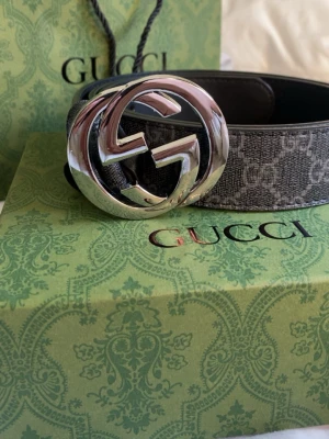 Gucci GG Supreme bälte med silverdetalj - Snyggt Gucci GG Supreme bälte i mörkgrått med klassiskt GG-mönster och stor silverfärgad GG-spänne. Perfekt accessoar för att lyfta vilken outfit som helst. Kommer med originalförpackning och kontrollkort.
