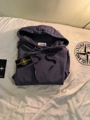 Stone Island hoodie - Snygg lila hoodie från Stone Island med klassisk huva och dragsko. Hoodien har en stor magficka och den ikoniska Stone Island-loggan på vänster ärm. Tillverkad i mjuk bomull för skön känsla och avslappnad passform. Perfekt för dig som gillar streetwear och vill sticka ut.