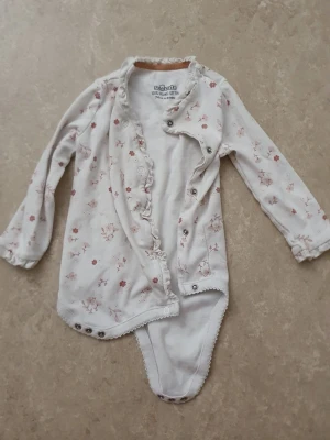 Babybody - Supersöt långärmad body från So cute i vit med små blommor i rosa och brunt. Volangkant längs knäppningen framtill och tryckknappar i grenen för enkel påklädning. Mjuk och skön bomull, perfekt för mysiga dagar.