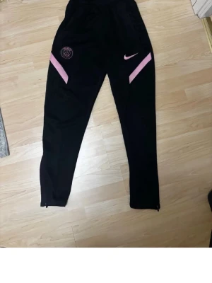 Svarta PSG träningsbyxor Nike - Säljer ett par svarta träningsbyxor från Nike med PSG-logga och rosa detaljer. Byxorna har smal passform, dragkedja vid benslut och är tillverkade i ett mjukt funktionsmaterial. Perfekta för fotbollsträning eller chill.