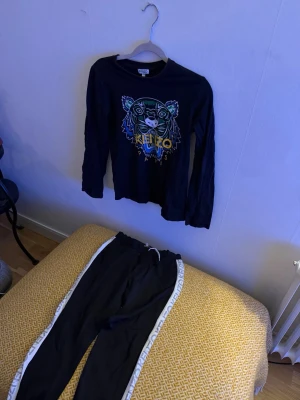 Kenzo set - Säljer ett par svarta mjukisbyxor från Kenzo med vita revärer längs sidorna. Byxorna har en avslappnad passform och resår i midjan för extra komfort. Även en kenzotröja. Perfekta för chill dagar eller när du vill ha en sportig look. 