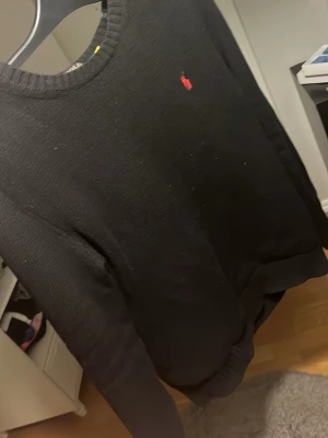 Svart tröja från Ralph Lauren - Klassisk svart tröja från Ralph Lauren med rund halsringning och den ikoniska röda logotypen broderad på bröstet. Tröjan har långa ärmar och ribbade muddar vid ärmslut och nederkant. Perfekt för en stilren och enkel look.