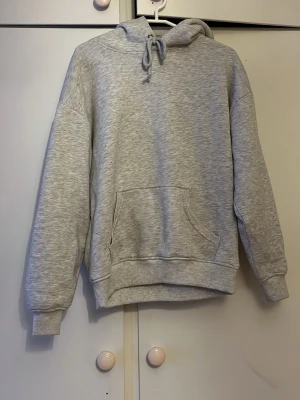 Ljusgrå hoodie med magficka - En stilren ljusgrå hoodie med huva och dragsko. Hoodien har en klassisk magficka och ribbade muddar vid ärmslut och nederkant. Perfekt för en avslappnad look och enkel att matcha med andra plagg.