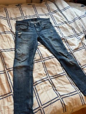 Replay anbass Blå slitna skinny jeans  - Säljer ett par blå skinny jeans från replay med slitningar och hål på låren och knäna. Jeansen har klassisk femficksmodell, låg midja och dragkedja. Perfekta för dig som gillar en avslappnad och trendig stil.