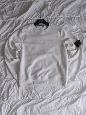 Vit sweatshirt  - Säljer en clean vit sweatshirt med klassisk rund hals och ikonisk logga på ärmen. Tröjan är i mjuk bomull och har ribbade muddar vid ärmslut och nederkant. Perfekt för dig som gillar stilrena plagg med streetwear-vibe.