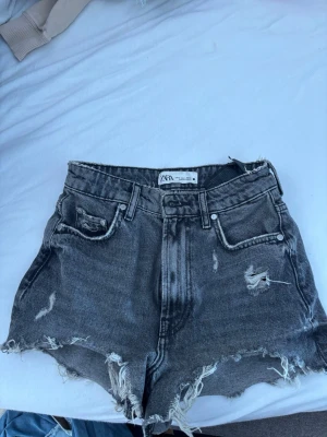 Gråa high waist jeansshorts Zara - Snygga gråa jeansshorts från Zara med hög midja och slitna detaljer. Shortsen har råa kanter, fem fickor och en klassisk knappgylf. Perfekta för en avslappnad och trendig look.