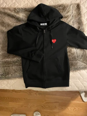 Svart hoodie Comme des Garçons Play - Svart hoodie från Comme des Garçons Play med dragkedja, huva och snörning. På bröstet sitter det ikoniska röda hjärtat med ögon. Hoodien har två fickor framtill och är tillverkad i mjuk bomull. Perfekt för en clean och stilren streetwear-look.