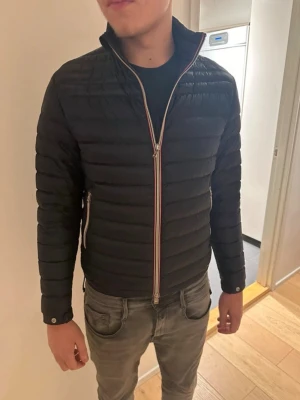 Mörkblå moncler dunjacka med randig dragkedja - Snygg mörkblå dunjacka med quiltad design och ståkrage. Jackan har en randig dragkedja framtill och sidofickor med dragkedja. Har ett litet håll i ena ärme.