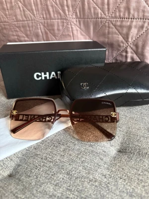 Bruna fyrkantiga Chanel solglasögon - Säljer ett par lyxiga solglasögon från Chanel med fyrkantiga, bruna tonade glas och breda skalmar med utskuren logga och guldfärgade detaljer. Kommer med originaletui i svart quiltat skinn och box. Perfekt accessoar för en trendig look.
