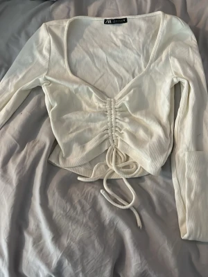 Vit ribbad topp från Zara - Trendig vit långärmad topp från Zara med ribbad struktur och djup urringning. Toppen har snörning och rynkdetalj framtill som ger en snygg passform. Perfekt att styla med jeans eller kjol för en nice look