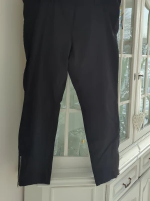 Svarta byxor från Imperia - Stilrena svarta byxor från Imperia med klassisk rak passform och bälteshällor. Byxorna har dragkedja vid benslut. Lite stretch. Dragkedja och knäppning i sidan. 