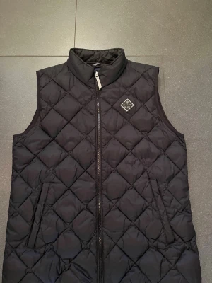 Svart quiltad dunväst från Gant - Snygg svart dunväst från Gant med quiltat mönster och hög krage. Västen har två fickor med dragkedja och en diskret logotyp på bröstet. Perfekt att slänga över en hoodie eller tröja för extra värme. Materialet är mjukt och lätt, vilket gör den enkel att bära.