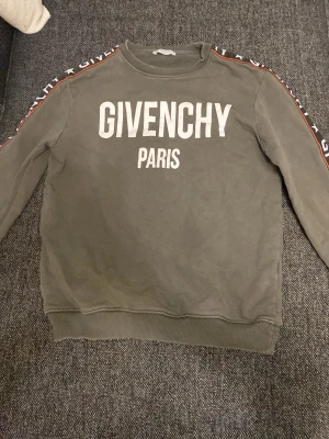 Grå sweatshirt från Givenchy - Säljer en grå sweatshirt från Givenchy med stor vit logga och texten 'PARIS' på bröstet. Tröjan har ribbade muddar och dekorativa band med Givenchy-tryck längs ärmarna. Perfekt för dig som gillar streetwear och exklusiva märken.