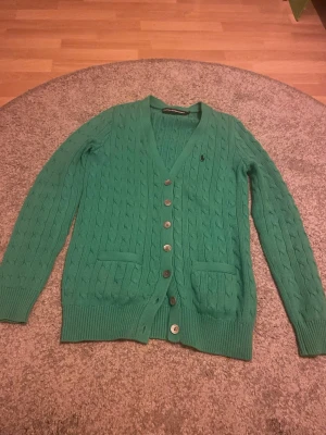 Grön kabelstickad kofta med knappar - Snygg grön kabelstickad kofta med v-ringning och sex knappar framtill. Två fickor framtill och ribbade muddar vid ärmslut och nederkant. Perfekt för lager på lager och ger en klassisk look.