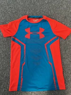 Under Armour röd och blå träningströja - Snygg kortärmad träningströja från Under Armour i rött och blått med stort logotryck på bröstet. Tillverkad i ett lätt och snabbtorkande funktionsmaterial som passar perfekt till gymmet eller löprundan.