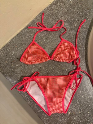 Rutig röd bikini med knyt - Söt bikini i rödvitt rutigt mönster med knyt i sidorna och runt nacken. Klassisk trekantsmodell med tunna band och fräscha färger, perfekt för stranden eller poolen. Aldrig använd, säljer för jag har för den inte kommer till användning 