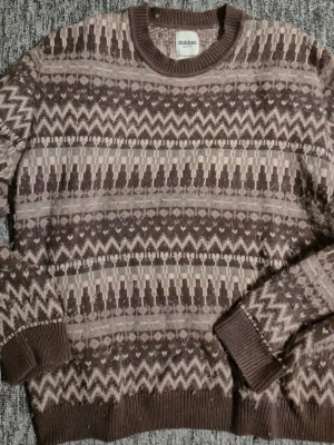 Brun mönstrad stickad tröja Dobber (MQ)  - Stickad tröja från Dobber (MQ) i bruna och beige toner med ett retroinspirerat mönster. Tröjan har rund halsringning, ribbade muddar och är långärmad. Perfekt för dig som gillar klassiska mönster och mjuka material. Varm tröja. Storlek XL, Finns i Hässelby 