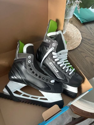 Bauer svarta hockeyskridskor - Säljer ett par Bauer hockeyskridskor i svart med vita och gröna detaljer. Skridskorna har vit sula, vita snören och grön insida. Yttermaterialet är i slitstark syntet med modern design och extra stöd runt ankeln. Perfekta för isträning,hockey eller bara ha kul på isen. De är också helt nya, aldrig använda. Köpte de förra året för detta år men märkte att jag tyvärr vuxit ur de. Paket och skydd finns även. Storlek 10 USA vilket motsvarar 43-44 EU storlek 