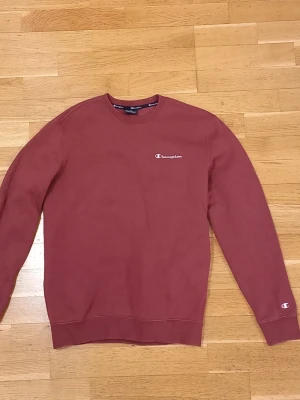 Vinröd sweatshirt från Champion - Säljer en vinröd sweatshirt från Champion med klassisk logga broderad på bröstet och ärmen. Tröjan har rund halsringning, ribbade muddar och är tillverkad i mjuk bomullsblandning. Perfekt för en avslappnad och stilren look.