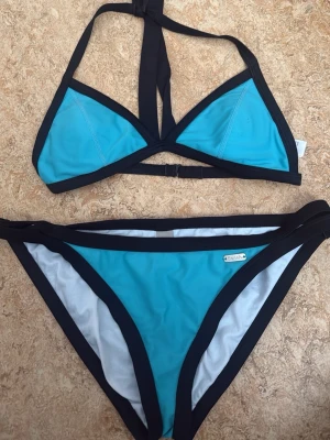 Blå bikini med svarta kanter från Tribord - Snygg blå trekantsbikini  med svarta kontrastkanter. Överdelen har knyt i nacken, och underdelen har dubbla svarta band i sidorna. Perfekt för strandhäng eller poolen! Dupe på Triangl bikini. Jättefint inför sommaren. Pris kan alltid diskuteras.