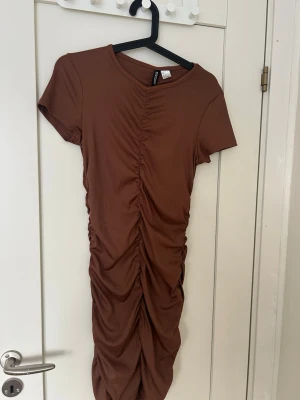 Brun ribbad kortklänning H&M - Kort brun klänning från H&M med ribbad struktur och rynkade detaljer längs hela framsidan. Klänningen har korta ärmar och en rund halsringning. Perfekt för dig som gillar en enkel men trendig stil.