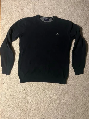 Mörkblå stickad tröja från Gant - Klassisk mörkblå stickad tröja från Gant med rund halsringning och diskret logga broderad på bröstet. Tröjan har långa ärmar och ribbade muddar vid ärmslut och nederkant. Perfekt basplagg i garderoben.