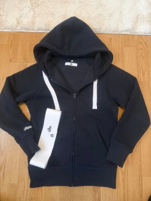 Russemerch Hoodie - Snygg marinblå hoodie från Russe merch med dragkedja framtill, stora fickor och vit dragsko i huvan. Tillverkad i mjukt bomullsmaterial och har ribbade muddar vid ärmar och nederkant. Perfekt för en avslappnad och cool stil.