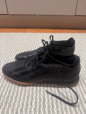 Svarta Adidas inomhusfotbollsskor - Säljer ett par svarta Adidas inomhusfotbollsskor med klassisk låg profil och gummisula i ljusbrunt. Skorna har snörning, diskreta svarta ränder på sidan och ovandel i syntetmaterial. Perfekta för futsal eller spel på inomhusplan.