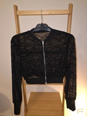 Kofta - Säljer en svart kofta med coola sammetsmönster i geometriska linjer. Jackan har dragkedja framtill, ribbade muddar vid ärmslut och midja samt rundad krage. Perfekt för dig som vill ha en edgy och unik look.