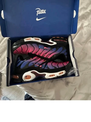 Nike x Patta Air Max Plus sneakers - Säljer ett par Nike x Patta Air Max Plus sneakers med en grym färgskiftning från rosa till blå, svarta detaljer och vita samt orangea inslag på sulan. Skorna har den klassiska TN-designen med vågiga linjer och synlig dämpning. Perfekta för dig som vill sticka ut.