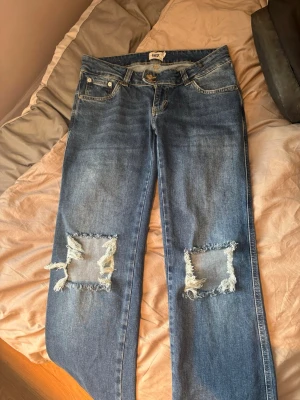 Blå slitna jeans med sopade knän - Säljer ett par blå jeans från Lager157 med raka ben och stora, fyrkantiga hål över båda knäna för en riktigt edgy look. Klassisk femficksmodell och knappgylf. Perfekta för dig som gillar streetstyle och vill sticka ut lite extra.