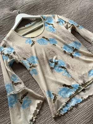 Beige blommig långärmad topp - Trendig beige topp med blåa blommor och långärmade trumpetärmar. Toppen har vågig kant nedtill och vid ärmslut, samt rund halsringning. Perfekt för dig som gillar en romantisk och bohemisk stil. Står ingen storlek men skulle säga att den passar S bäst 