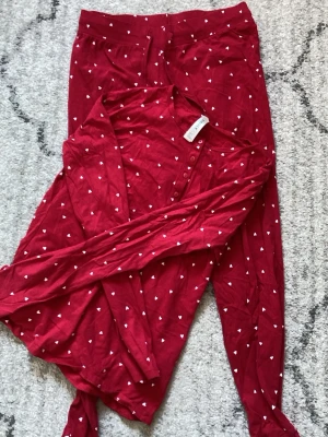 Röd pyjamas med hjärtmönster från Lindex - Mysig röd pyjamas från Lindex med små vita hjärtan över hela setet. Tröjan har långa ärmar och knappar vid halsen, byxorna har resår i midjan. Perfekt för chillkvällar hemma. Tillverkad i mjukt bomullsmaterial. Inte använt bara tvättat. Original pris 300kr