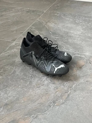 Svarta Puma fotbollsskor med vita detaljer - Säljer ett par svarta fotbollsskor från Puma med vita detaljer och logga. Skorna har snörning, strumpliknande skaft och dobbar för bra grepp på planen. Snygg design med grafiska mönster på sidorna och tåpartiet. Perfekta för dig som vill sticka ut på fotbollsplanen. Hör av er vid funderingar