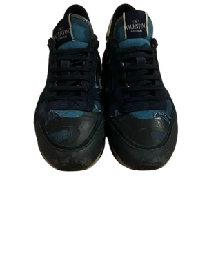 Valentino Garavani blå sneakers - Snygga sneakers från Valentino Garavani i blått och svart med kamouflagemönster. Skorna har klassisk låg profil, svarta snören och detaljer i mocka och textil. Perfekta för dig som vill ha en unik och trendig look.