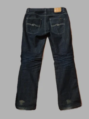 Nudie Jeans - Säljer ett par mörkblå jeans från Nudie Jeans 2001 med klassisk femficksmodell och kontrastsömmar. Jeansen har raka ben, snygga slitningar framtill och broderade bakfickor. Tillverkade i kraftigt denimtyg som ger en cool och tidlös look.